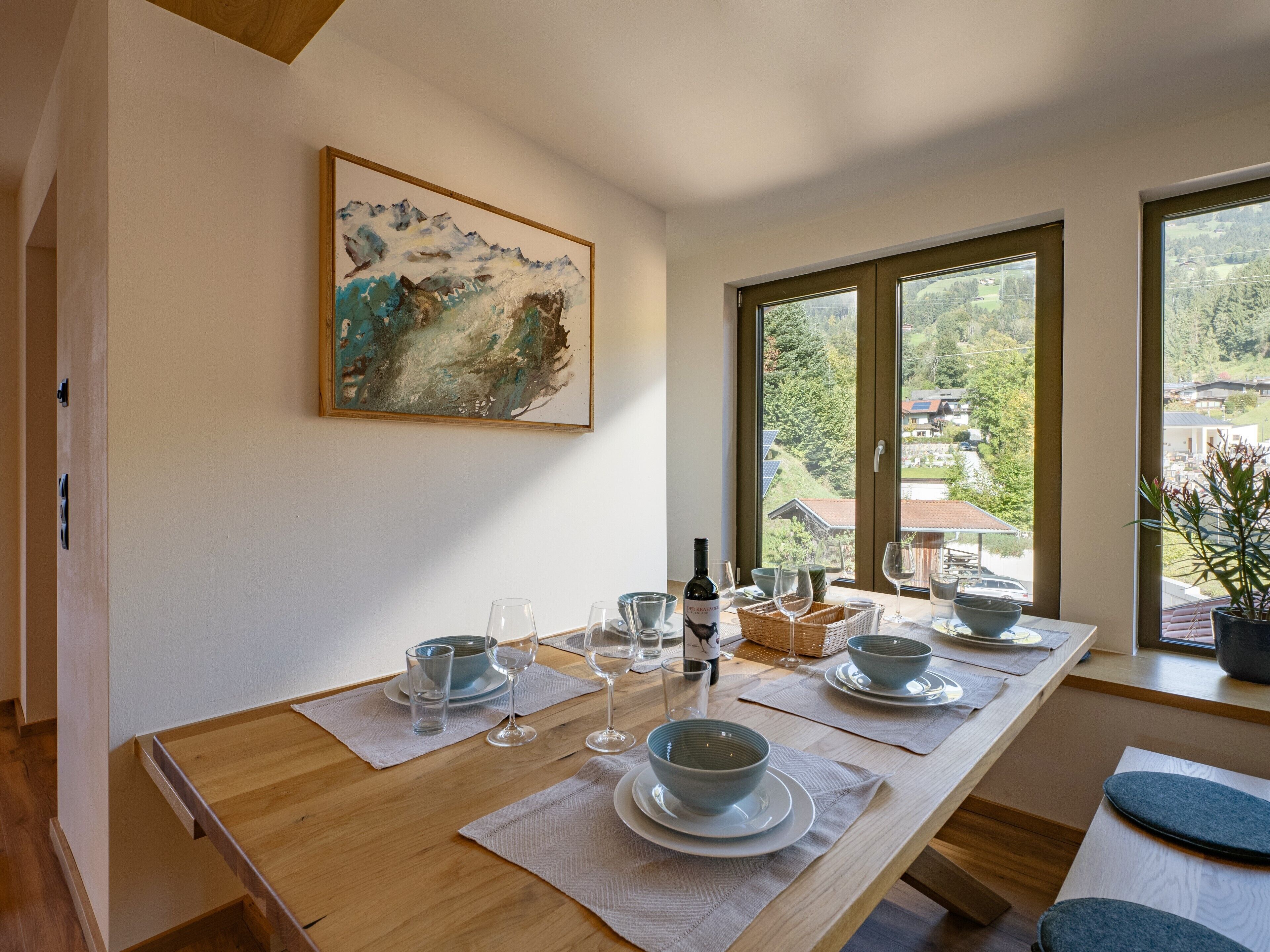 A dining area with a wooden table set for four, a painting on the wall, and a view of a village through large windows.