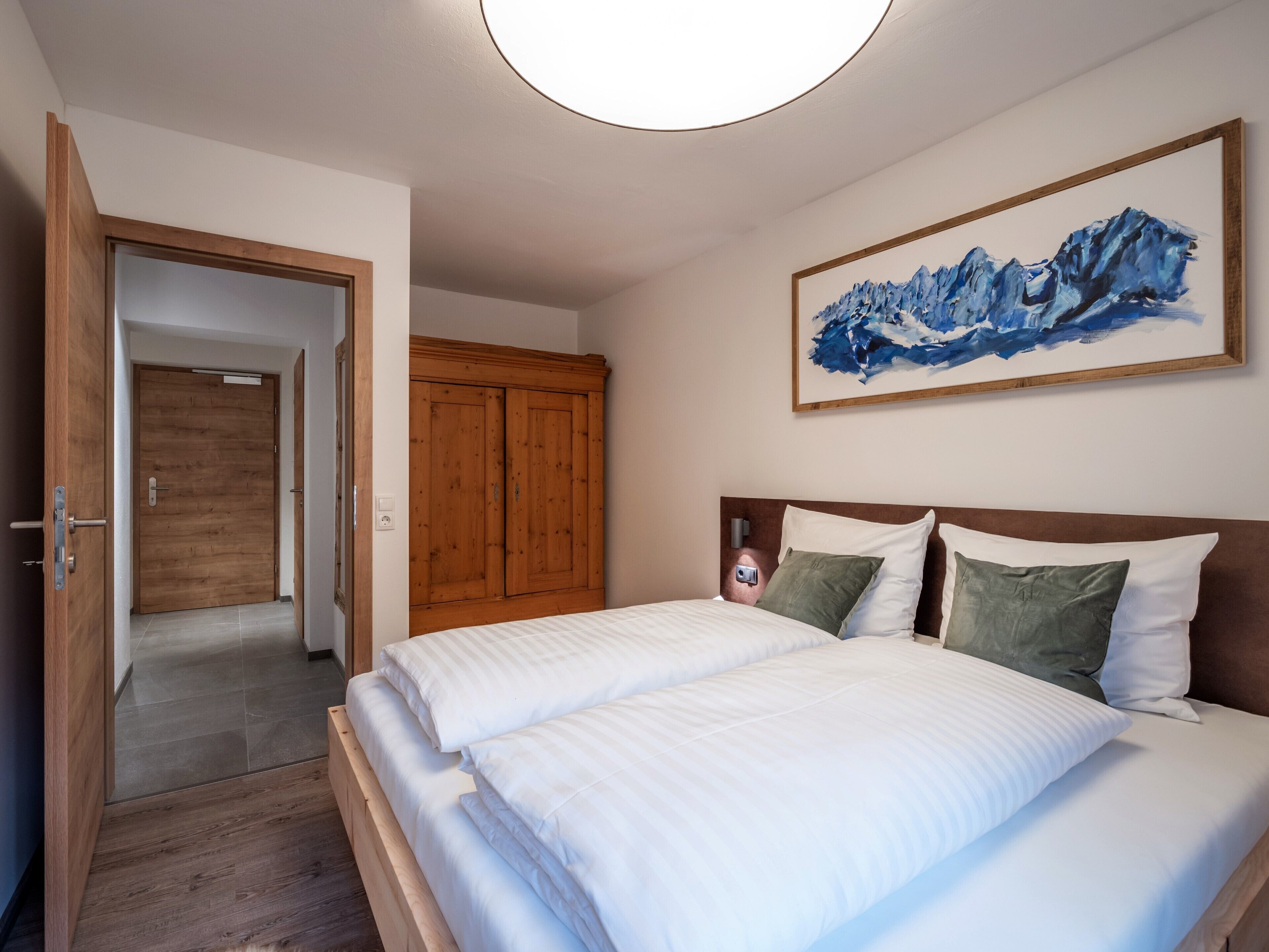 A bedroom with a wooden wardrobe, a bed with white bedding, and a painting of mountains on the wall.