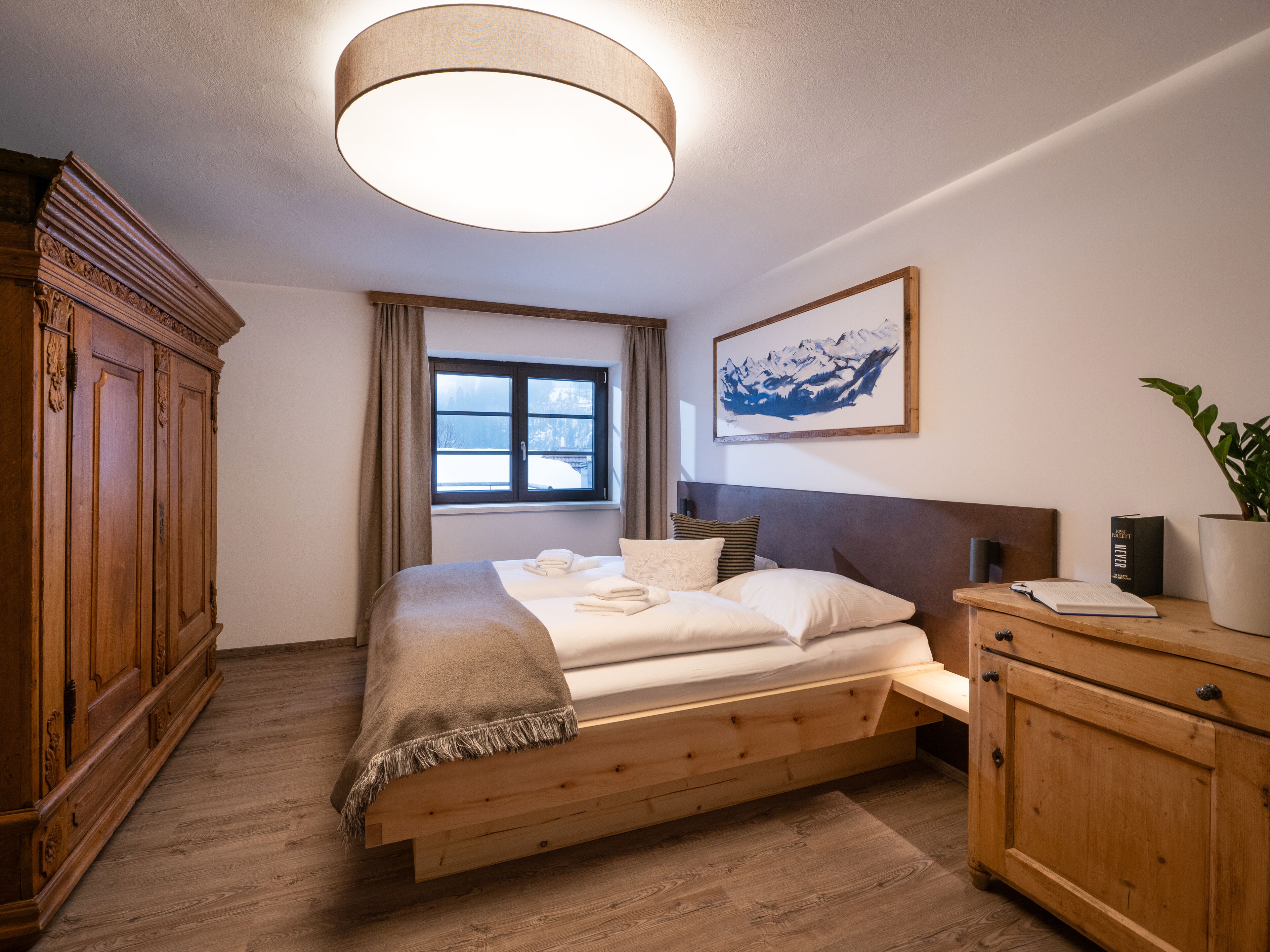 A bedroom with a wooden bed, a nightstand, a large window, a painting, and a wardrobe.
