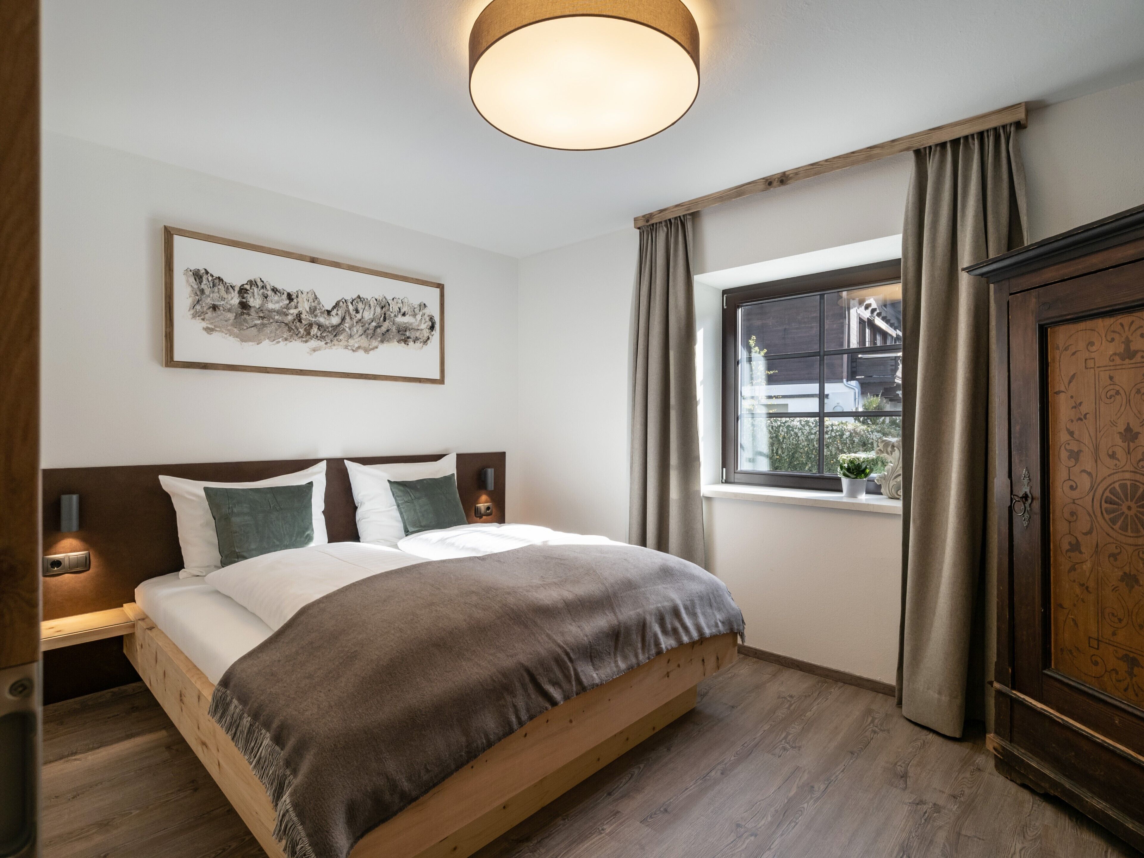 A bedroom with a wooden bed, a bedside table with a lamp, a window with curtains, and a wall-mounted artwork.