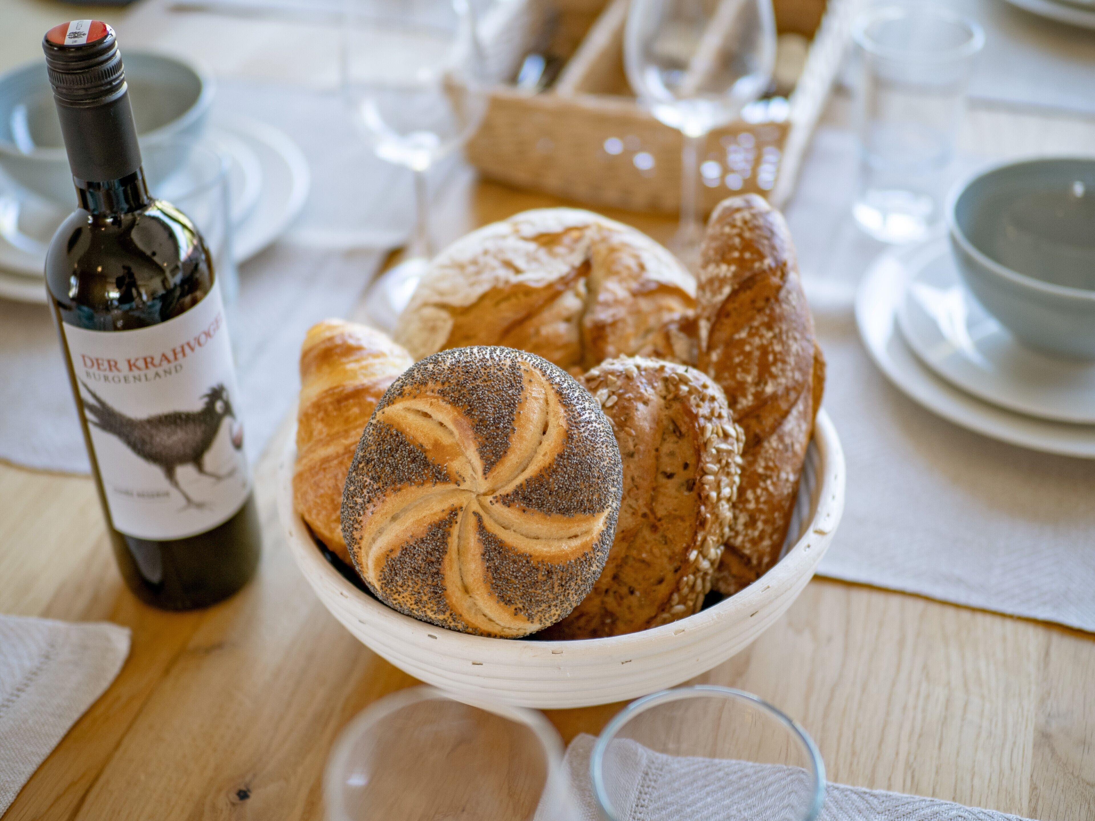 A table with a bowl of assorted bread, a bottle of wine, and a glass.