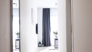 Ein Korridor mit Holzboden, einem Spiegel links, einer Tür rechts und einem Fenster mit Vorhang am Ende.