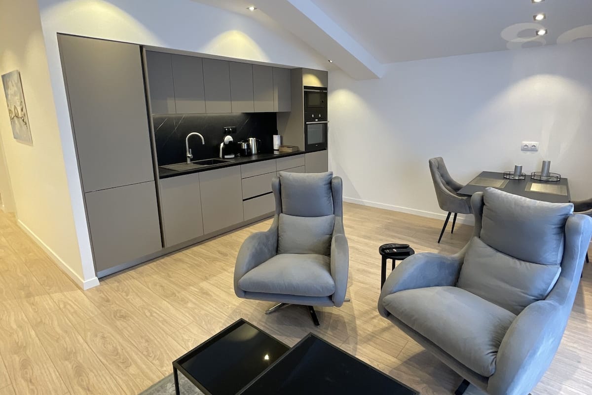 A modern living room with grey sofas, a black coffee table, and a kitchen area with grey cabinets and a black countertop.