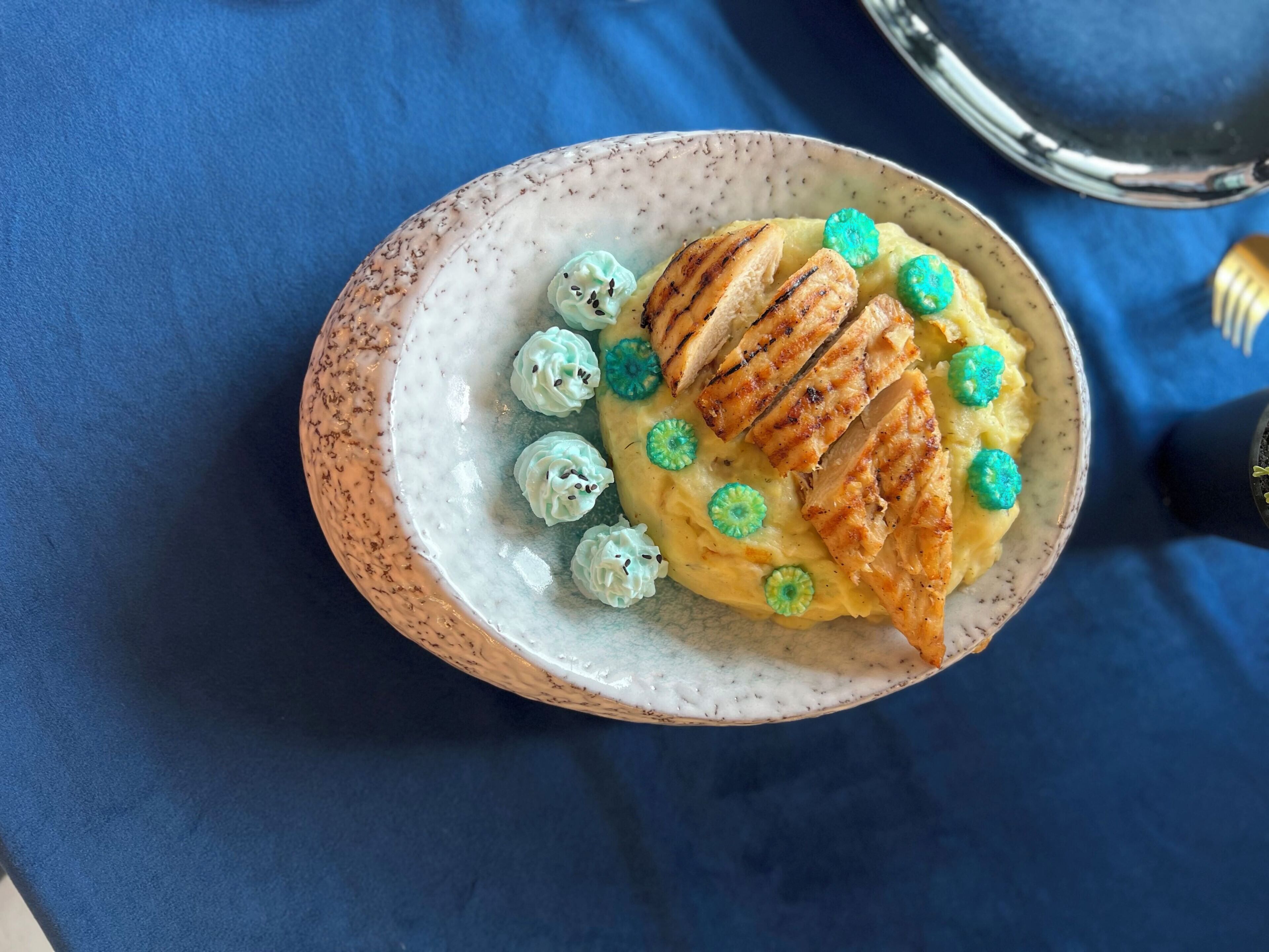 A bowl of food with grilled chicken and a creamy base, garnished with small green and blue items.