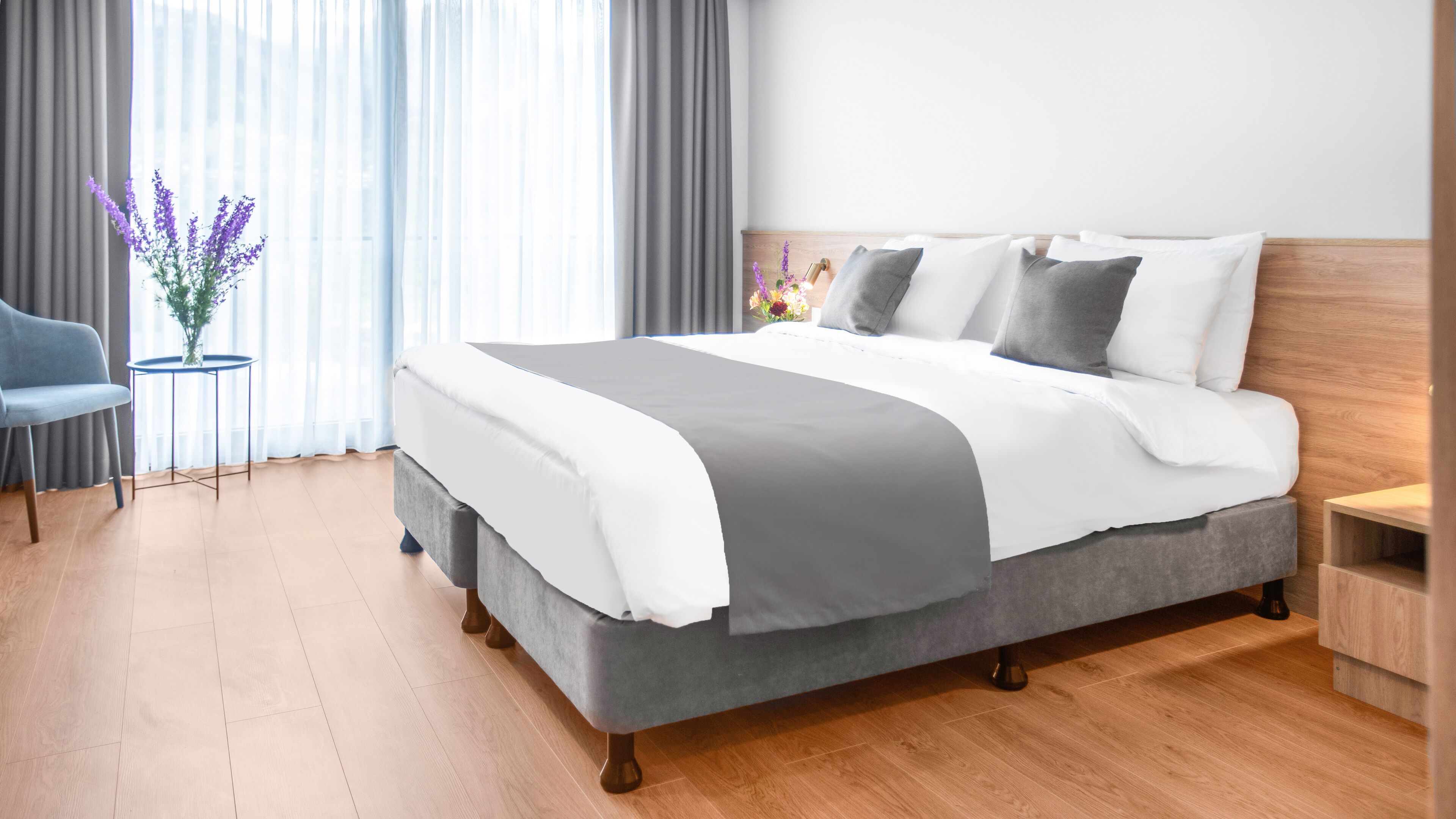 Una habitación de hotel moderna con una cama grande, ropa de cama gris y blanca, una mesita redonda con un jarrón de flores y un sillón azul.
