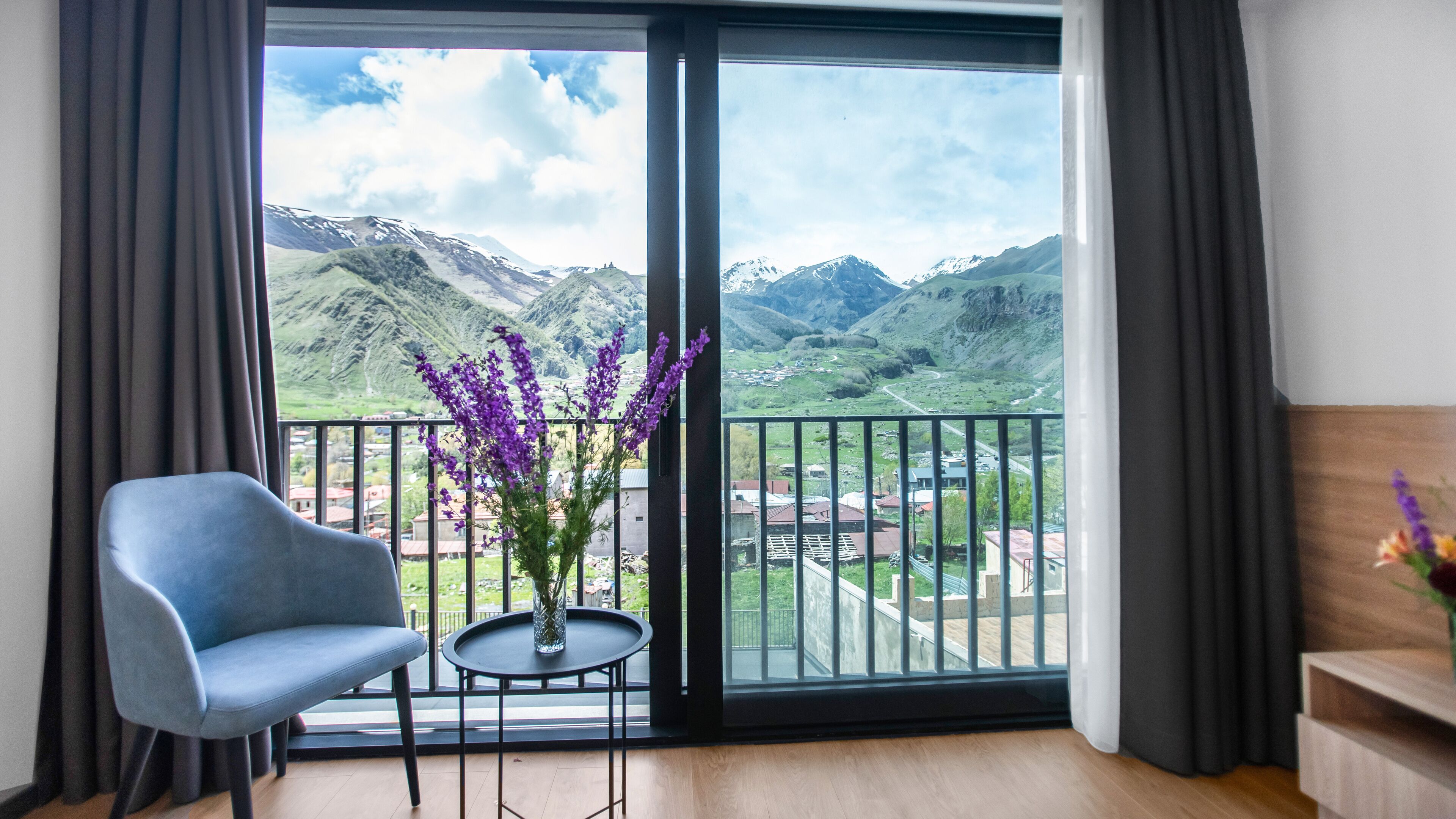 Una habitación con un ventanal que ofrece vistas a la montaña, una silla azul, una mesita con un florero y un armario de madera.