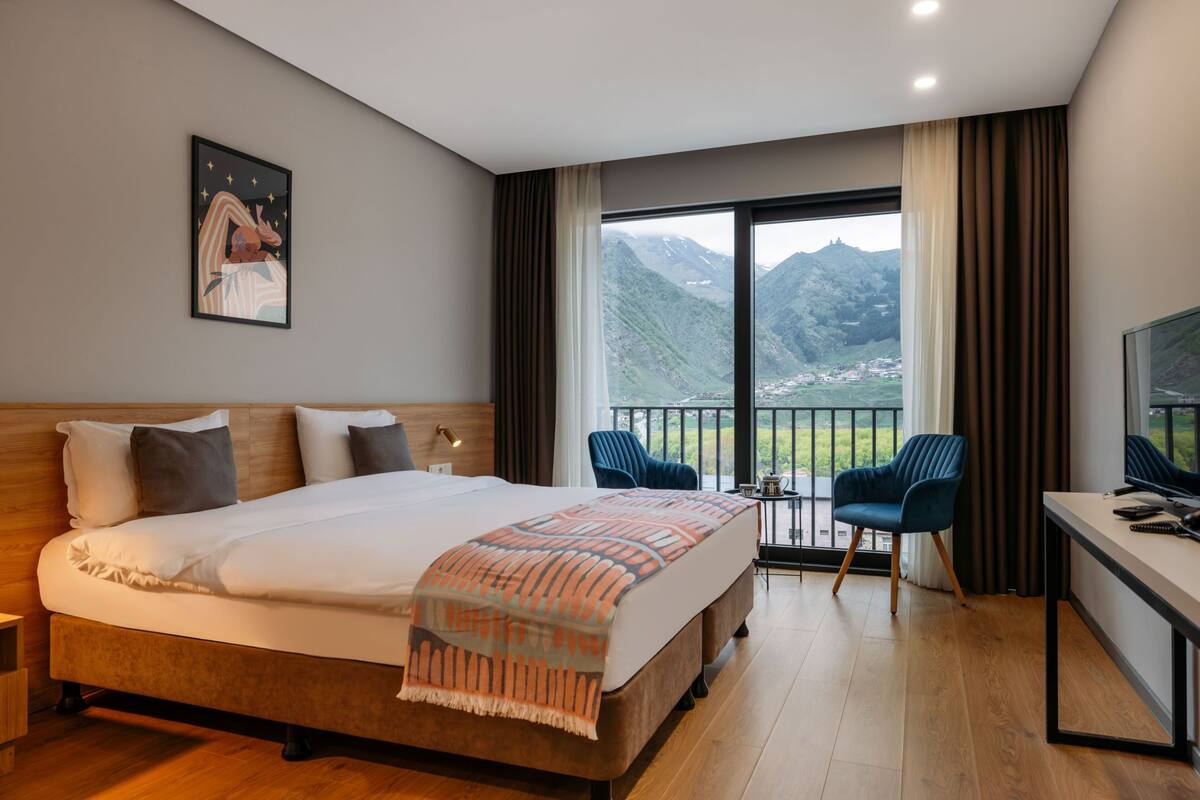 Habitación de hotel con una cama grande, un escritorio, una silla y un balcón con vista a las montañas.