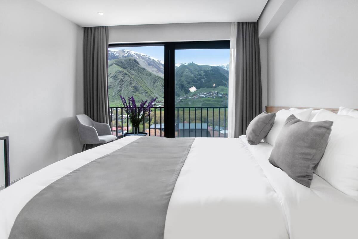 Habitación de hotel moderna con una cama grande, un balcón y vistas a la montaña, y un florero con flores moradas.
