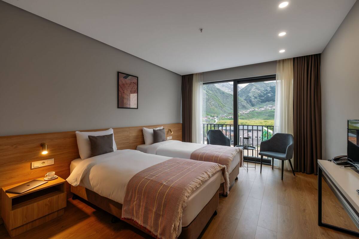 Habitación de hotel con dos camas, un escritorio con computadora, una silla y balcón con vista a la montaña.