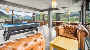 A pool table in a room with leather sofas and a wooden table, large windows with mountain views, and hanging pendant lights. - Hotel Memoir Kazbegi (Kazbegi)