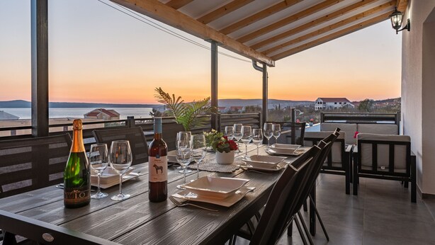 A dining table set for two with a bottle of wine, glasses, and a small plant. The table is outdoors with a view of the sea and buildings in the distance. - Villa Sea Pearl (Jasenice)