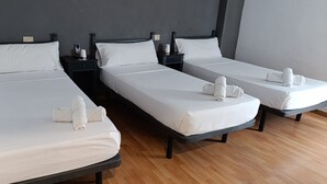 Habitación de hotel con tres camas individuales, cada una con ropa blanca y toallas. Mesitas de noche con lámparas y un enchufe. El suelo es de madera y las paredes son oscuras.