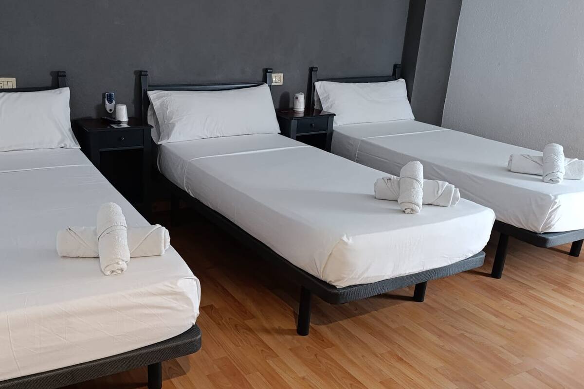 Habitación de hotel con tres camas individuales, cada una con ropa blanca y toallas. Mesitas de noche con lámparas y un enchufe. El suelo es de madera y las paredes son oscuras.