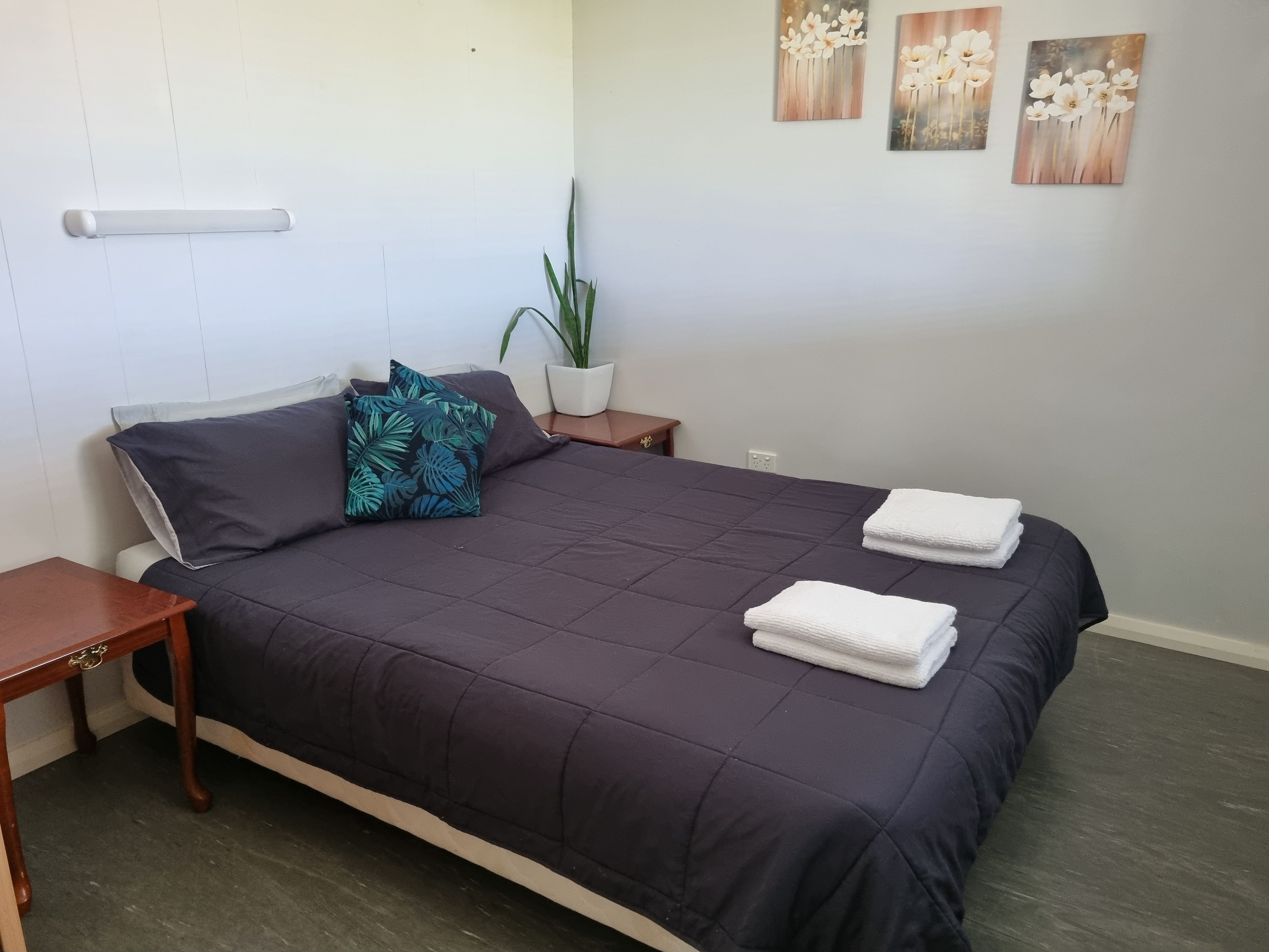 Ein ordentlich gemachtes Bett mit einer dunkelgrünen Tagesdecke und zwei Kissen, ein kleiner Nachttisch mit Lampe, ein Topfpflanze und drei an der Wand hängende Blumenbilder.