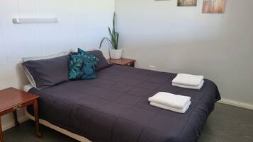 Ein ordentlich gemachtes Bett mit einer dunkelgrünen Tagesdecke und zwei Kissen, ein kleiner Nachttisch mit Lampe, ein Topfpflanze und drei an der Wand hängende Blumenbilder.