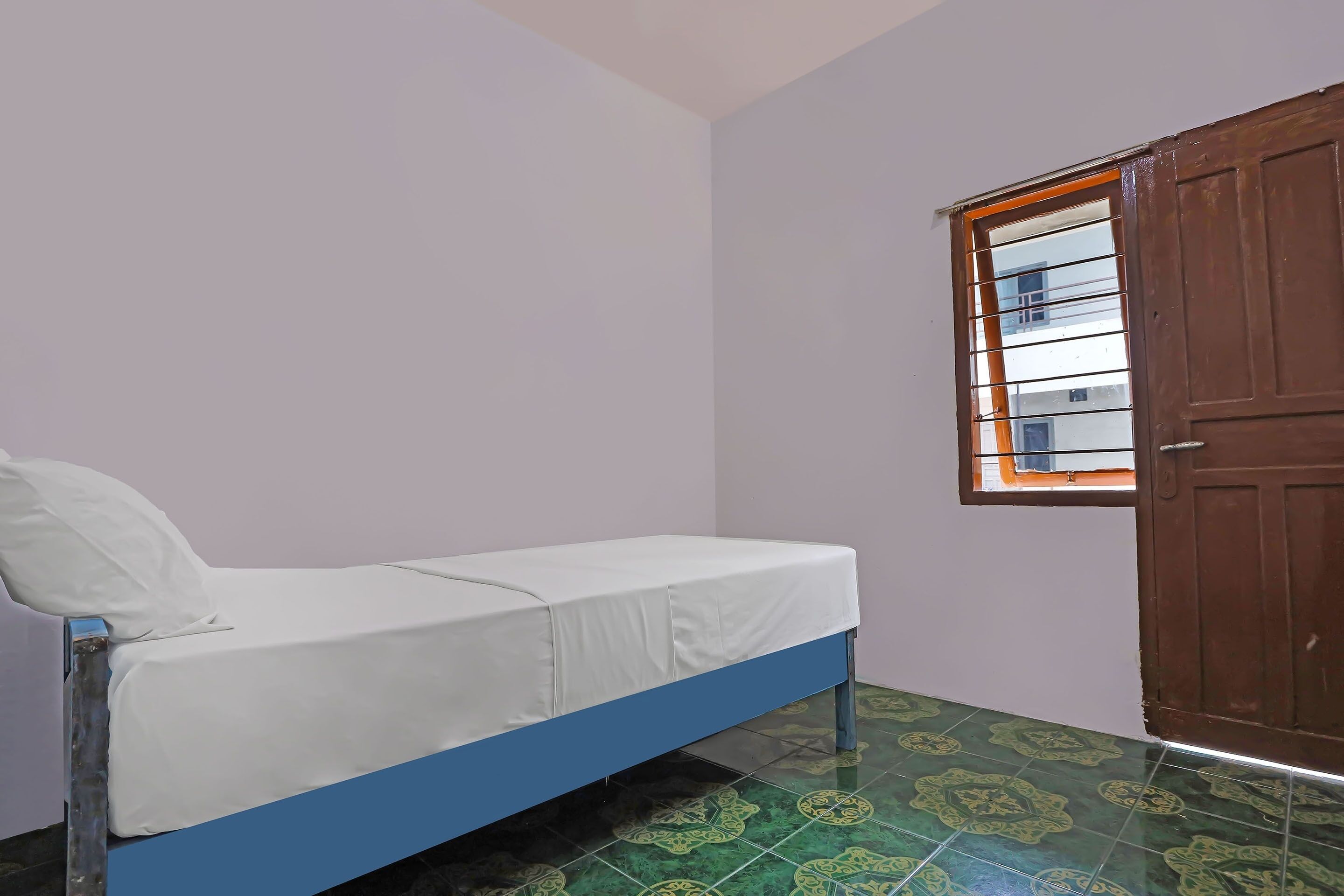 A small, single-bed room with a blue frame, white bedding, a window with orange blinds, and a green patterned floor.