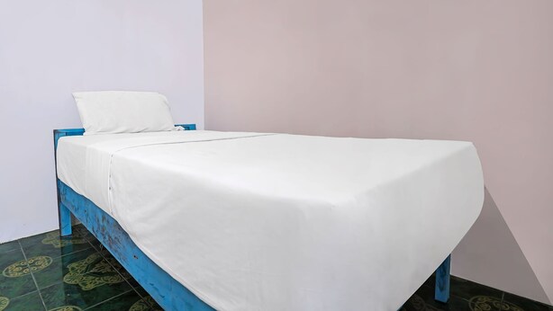 Uma cama de solteiro com lençol branco em quarto com estrutura azul e chão estampado.