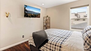 A bedroom with a bed, a mounted TV, a wall-mounted shelf with a plant, and a window with a view of a building. - Bright, Beautiful & Dog Friendly Steps to Old Town (Fort Collins)