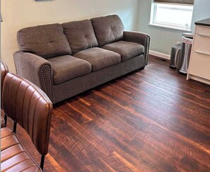 A room with a brown leather sofa, a wooden floor, and a window with blinds. - Bright, Beautiful & Dog Friendly Steps to Old Town (Fort Collins)