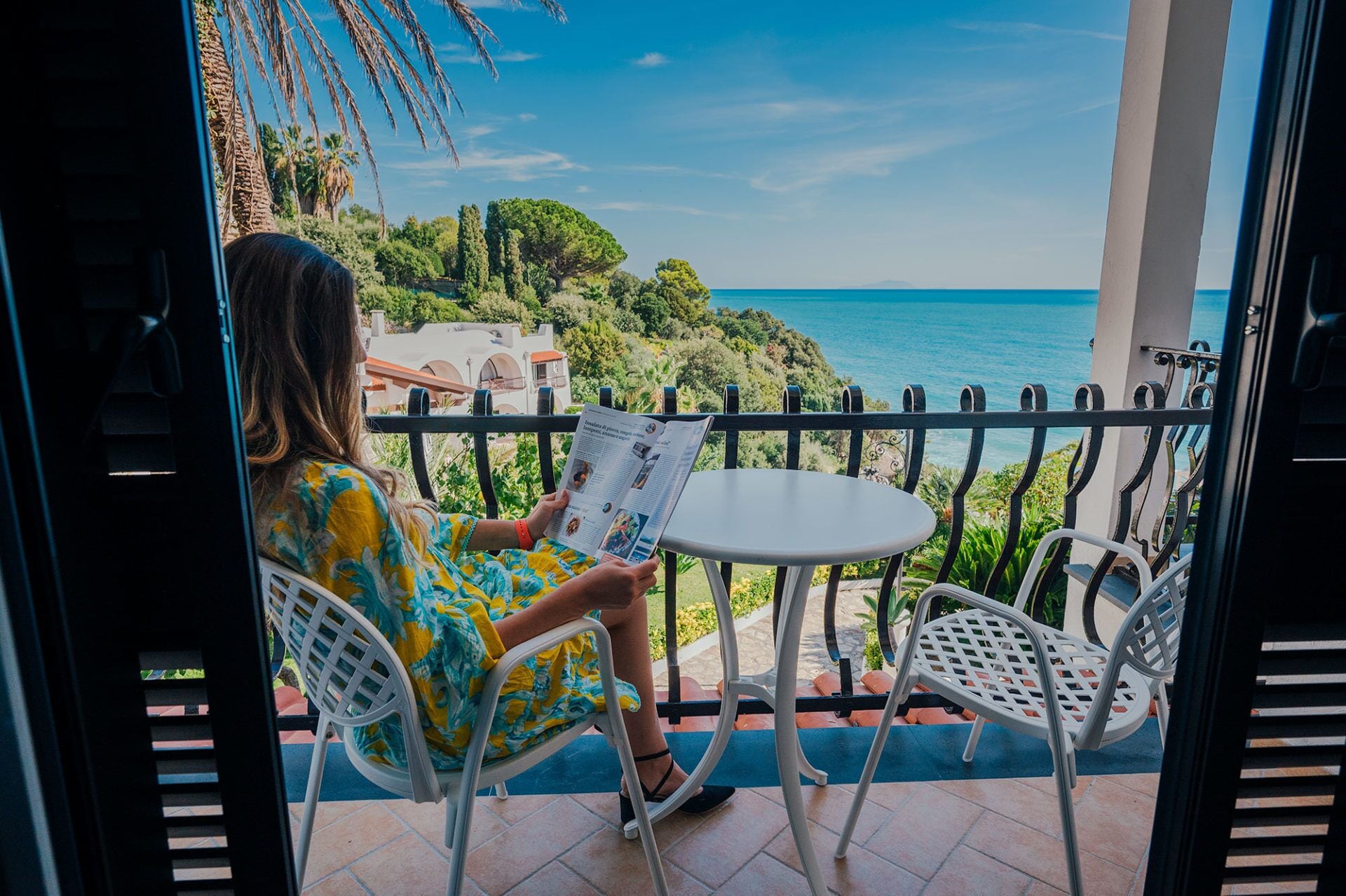 A person reading a magazine on a balcony with a view of the sea and greenery.