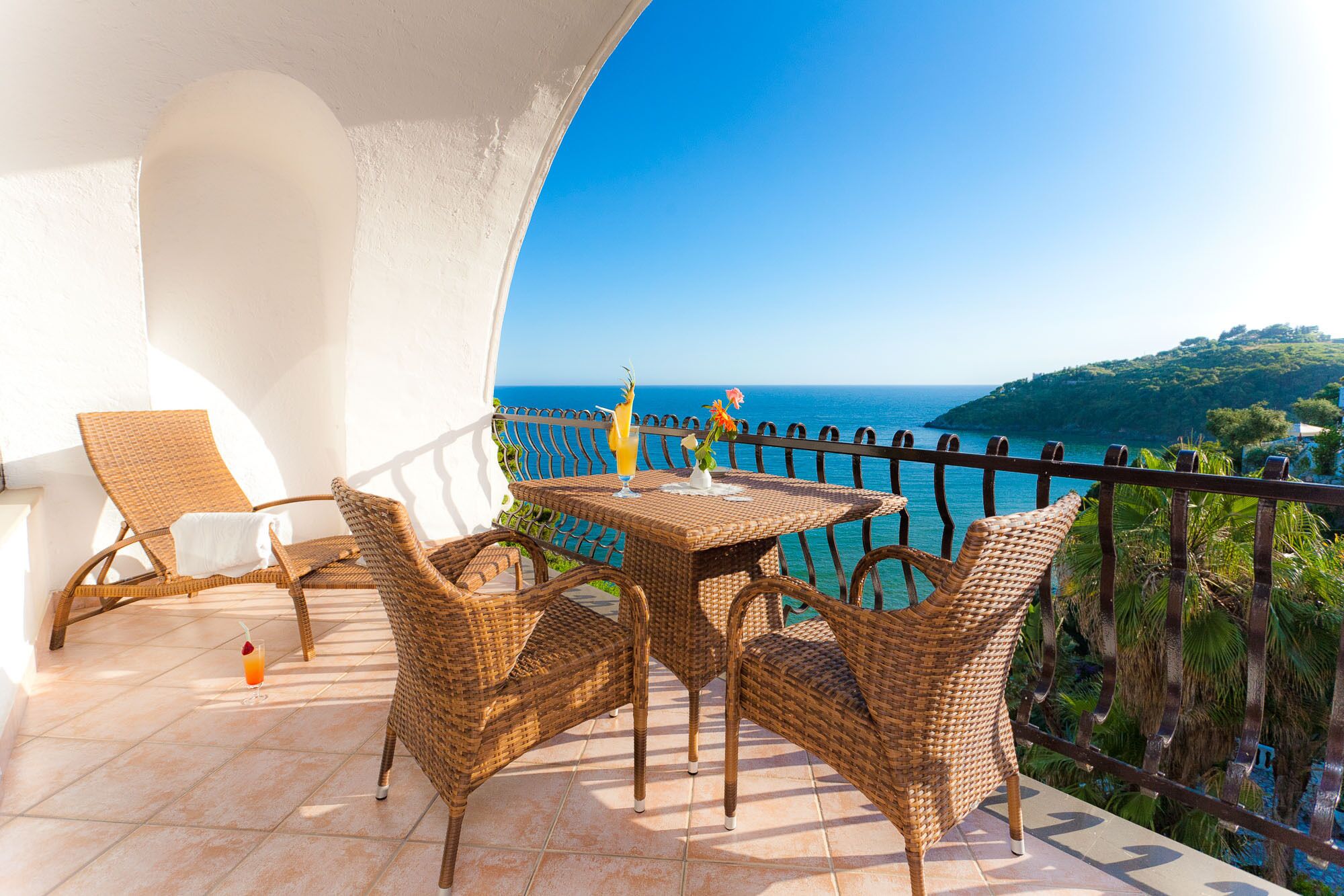 A balcony with wicker furniture, a table, and a drink, overlooking a coastal view.