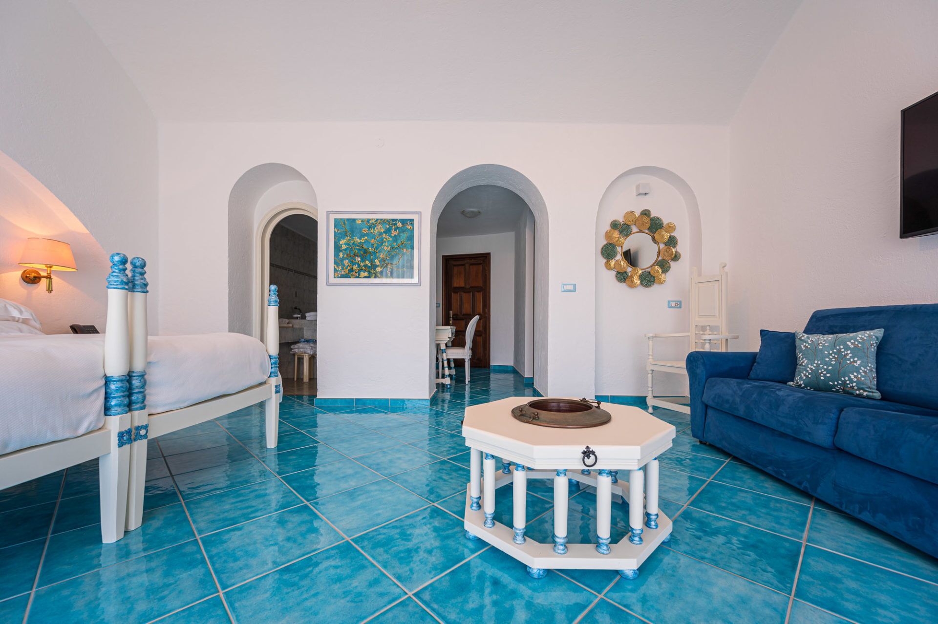 A hotel room with a bed, a blue sofa, a small table with a bowl, and a painting on the wall.