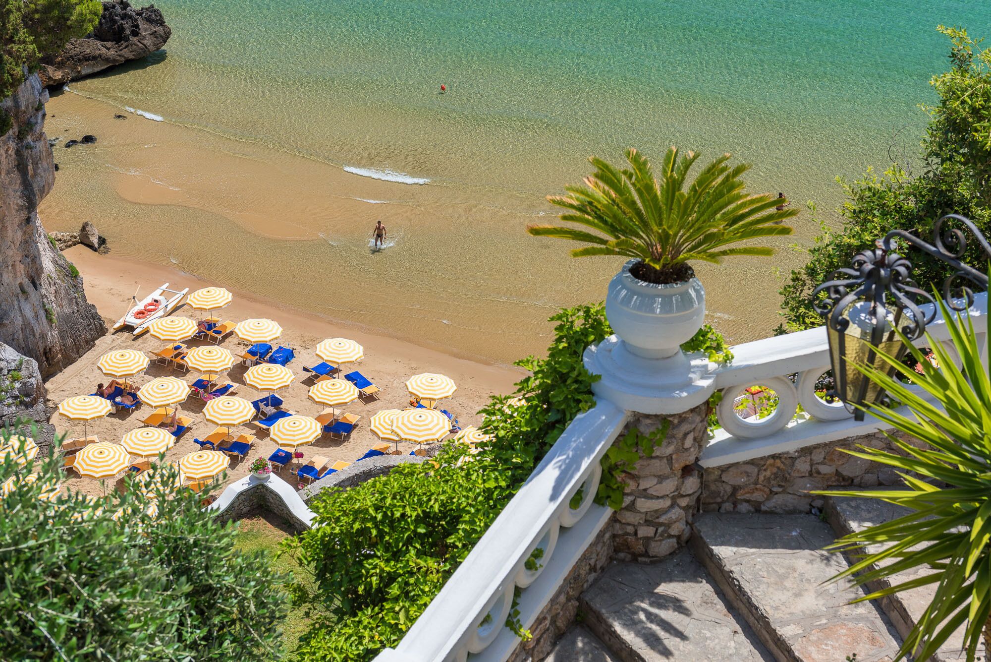 A beach with clear turquoise water, a stone staircase leading down, and numerous yellow and blue umbrellas and lounge chairs.