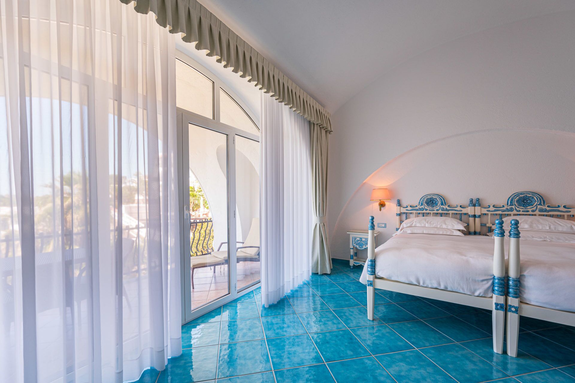 A bedroom with a bed, a nightstand, a lamp, and a view of a balcony.