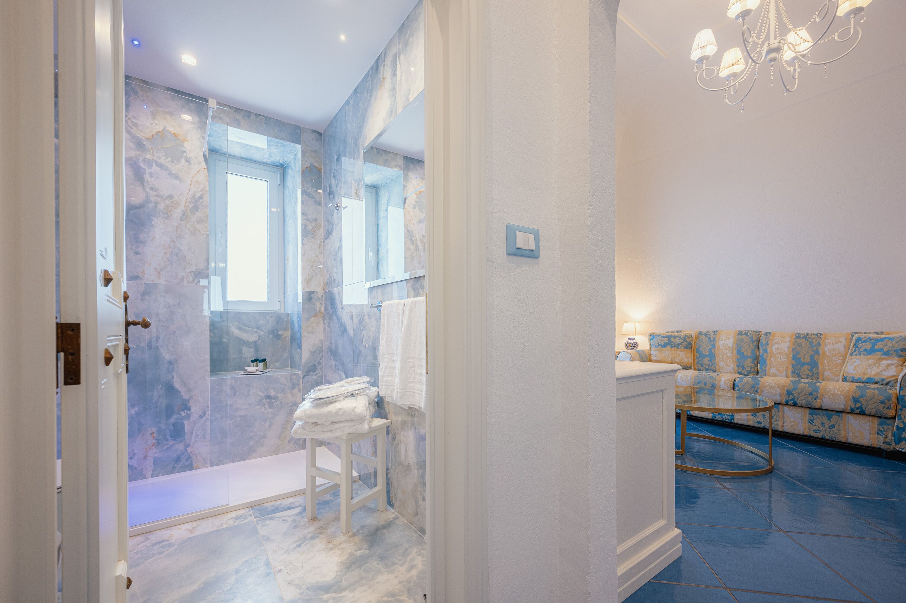 A bathroom with marble walls and a shower, a white sink, and a blue patterned sofa in the adjacent living area.