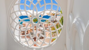 Une fenêtre circulaire ornée d’un motif géométrique, donnant sur un patio aménagé avec du mobilier d’extérieur et surplombant un ciel bleu.