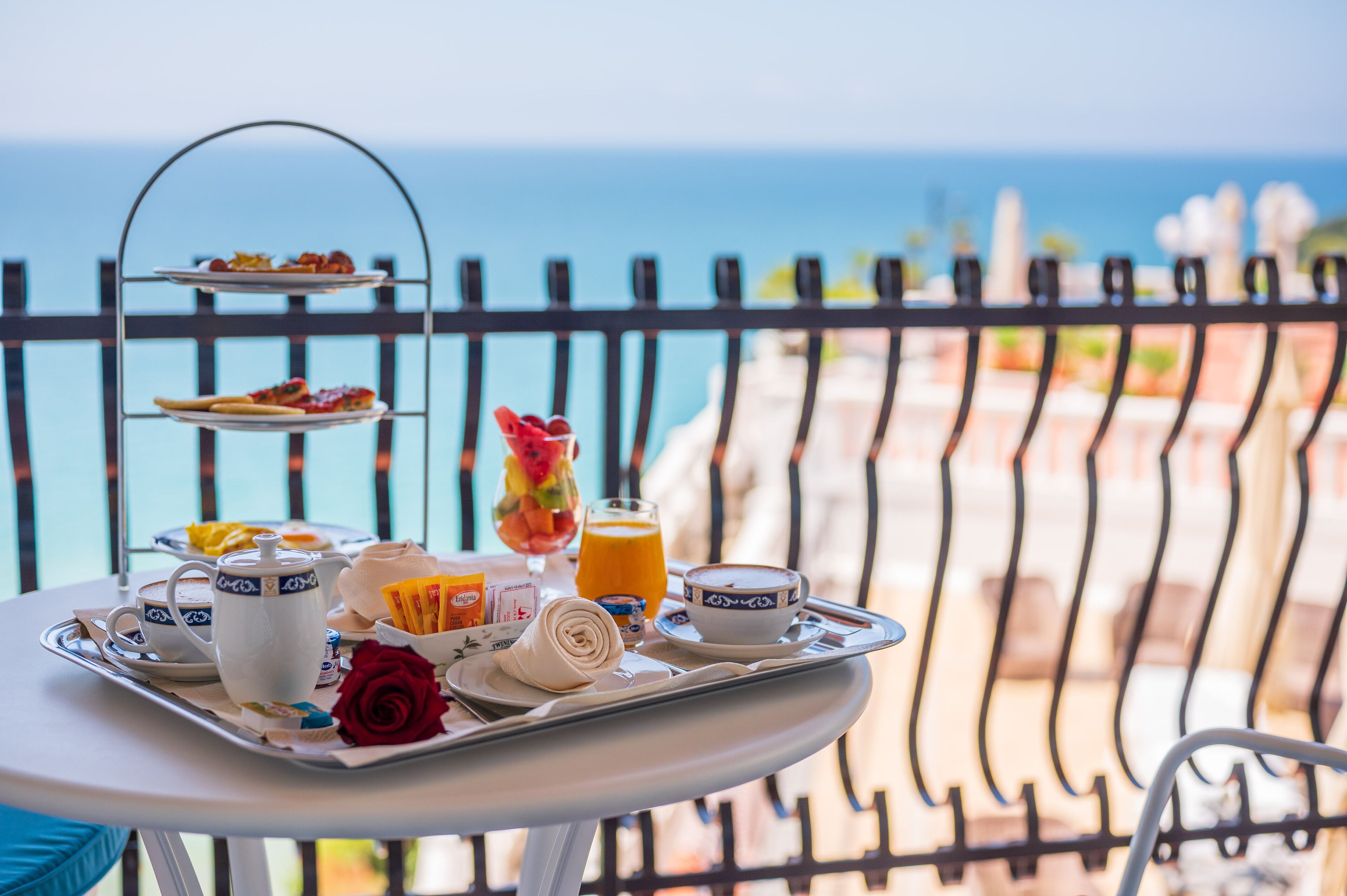 A breakfast tray with a teapot, cups, a bowl, a glass of orange juice, a rolled pastry, and a rose on a balcony overlooking the sea.