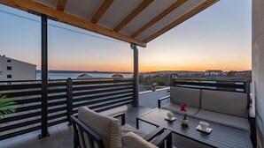 Ein Balkon mit Blick auf das Meer bei Sonnenuntergang, ein Tisch mit Tasse und Untertasse sowie eine Couch mit Kissen.