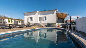 Ein Schwimmbecken mit Liegestühlen, ein weißes Gebäude mit einer überdachten Terrasse und einem strahlend blauen Himmel.