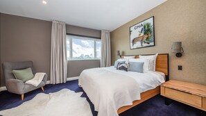 Une chambre d’hôtel avec un grand lit, une chaise, une table de chevet, une fenêtre avec des rideaux et une lampe fixée au mur.