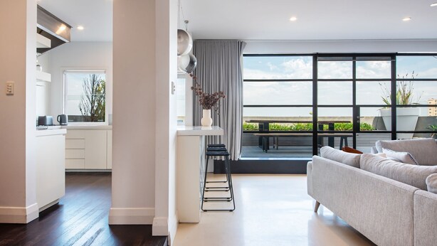 A modern living room with a large window, a sofa, and a kitchen area. - Pet Friendly - Penthouse with City Views on Chapel (St Kilda)