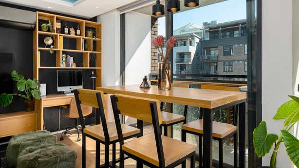 A modern dining area with a wooden table and chairs, a bookshelf with plants, and a view of buildings through large windows. - The Iron Works 611 (Cape Town)