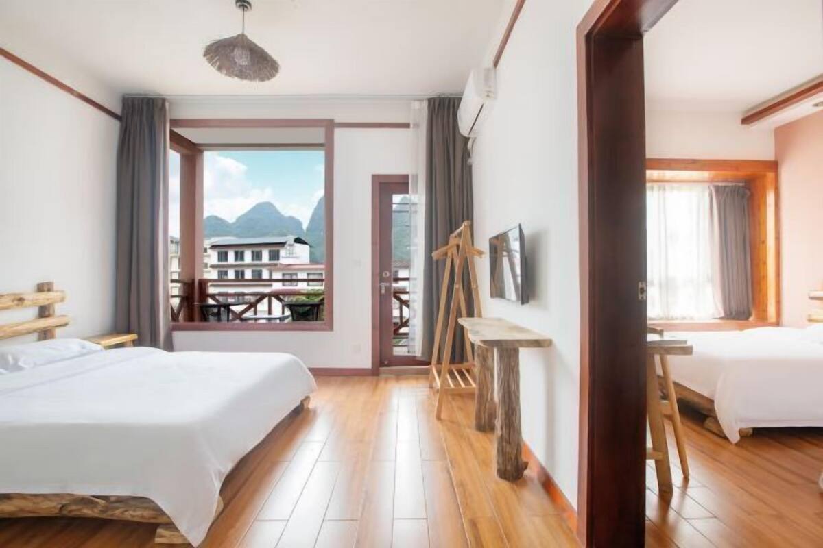 A hotel room with two beds, a wooden table, and a view of mountains through a large window.