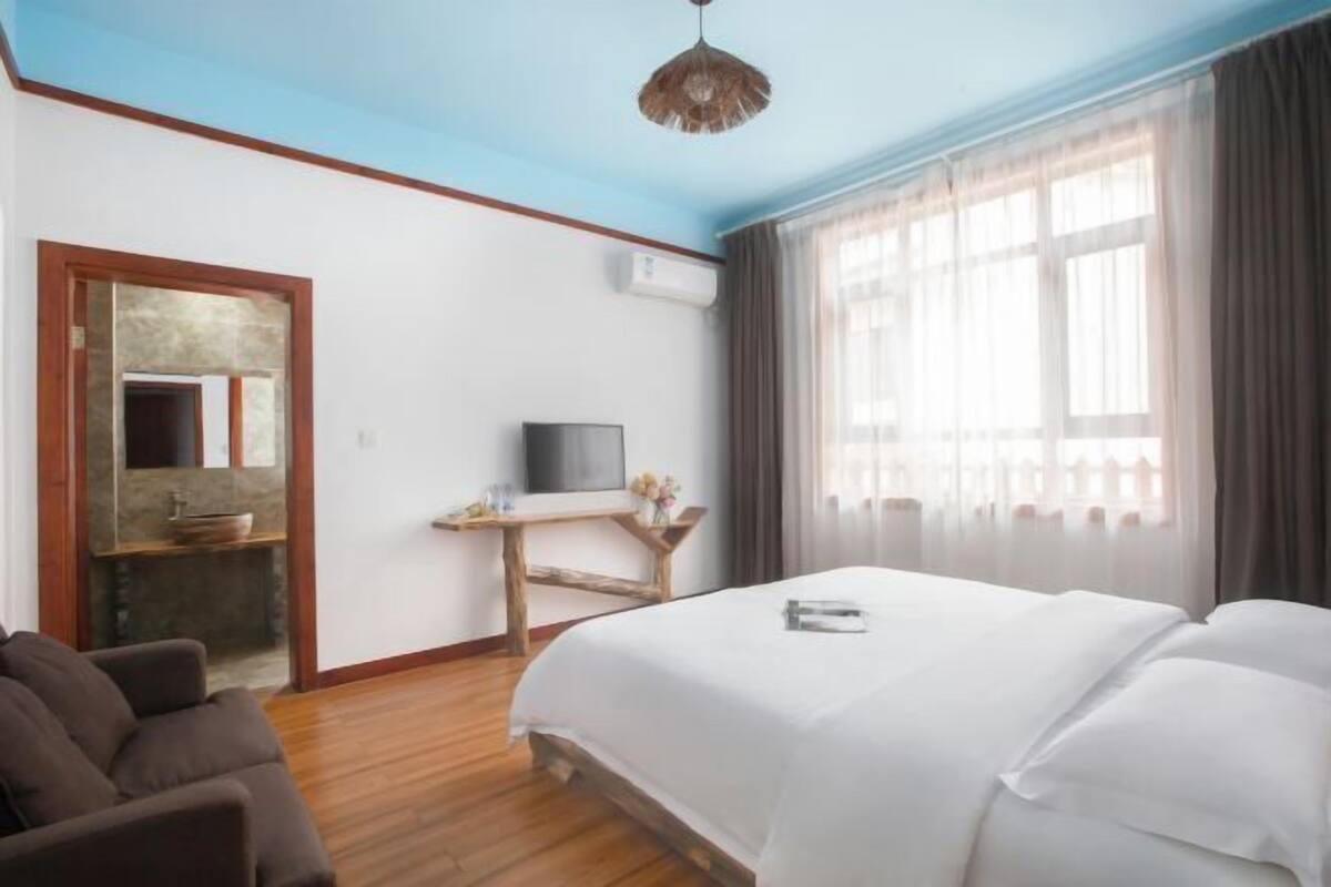 A hotel room with a bed, a TV, a wooden desk, a sofa, and a window with curtains.