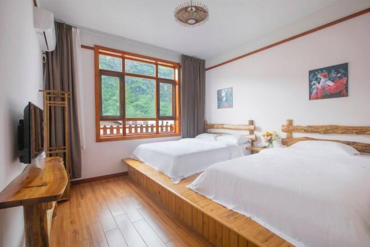 A room with two beds, a wooden bench, a TV, and a window with a view of greenery.