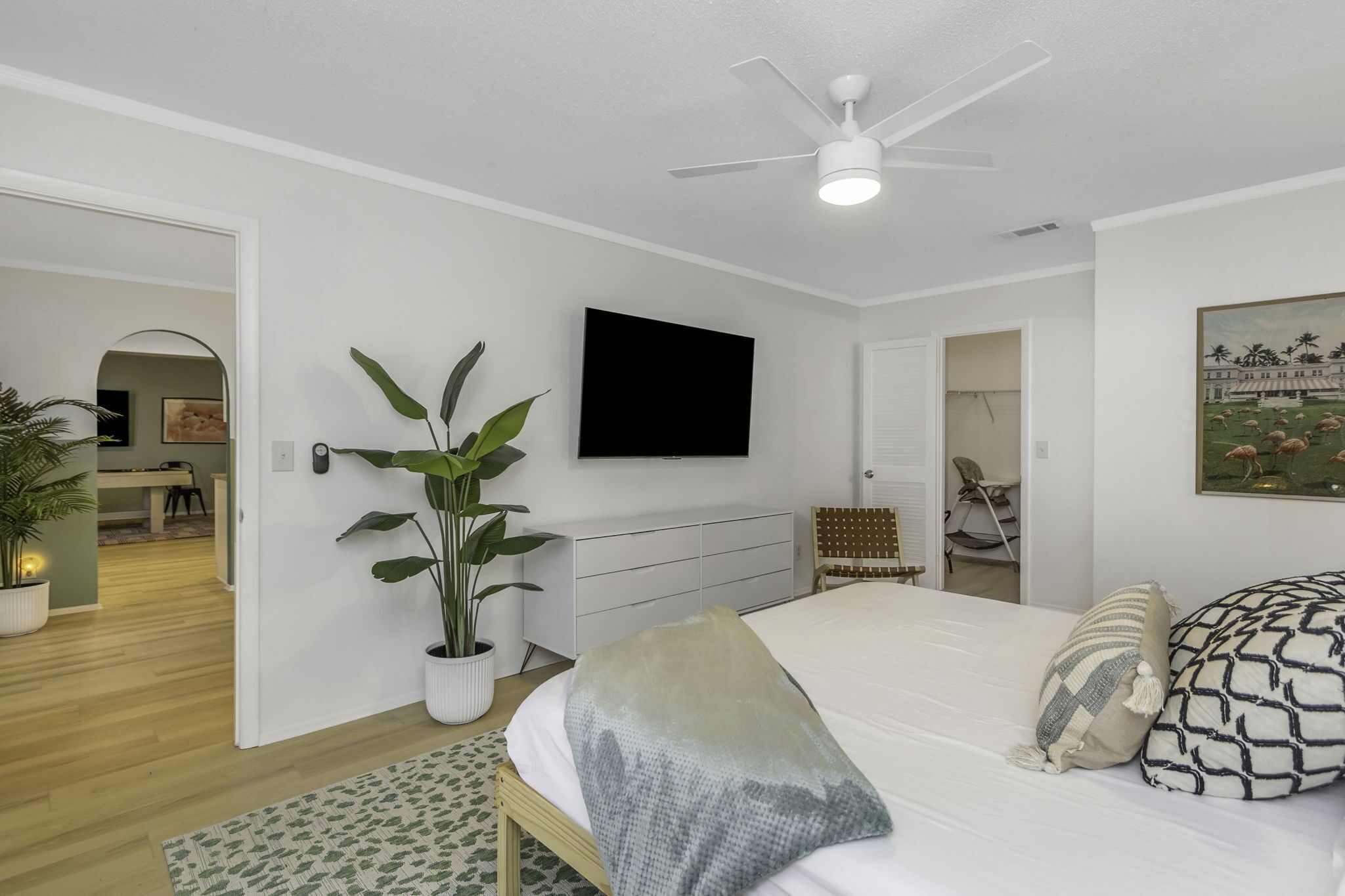 Un dormitorio con una cama, ventilador de techo, televisor, planta en maceta y un cuadro en la pared.