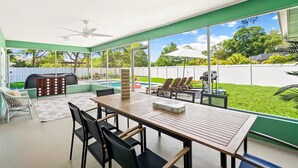 Un porche techado con una mesa y sillas, un ventilador de techo y vistas a un patio trasero con piscina y sillas de jardín.