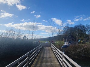 Eine Holzbrücke mit Geländern, die zu einer grünen Wiese mit Bäumen und blauem Himmel führt.