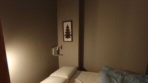Een kleine, zwak verlichte kamer met een enkel bed, een wandlamp en een lijst met een afbeelding van een boom.