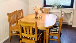 Un comedor con una mesa redonda de madera, seis sillas de madera, una lámpara colgante, una ventana con persianas y un radiador debajo.