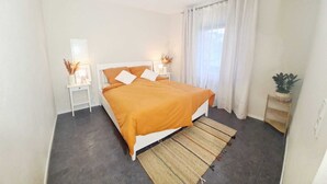 Un dormitorio con cama, mesitas de noche, lámpara, planta y ventana con cortinas.
