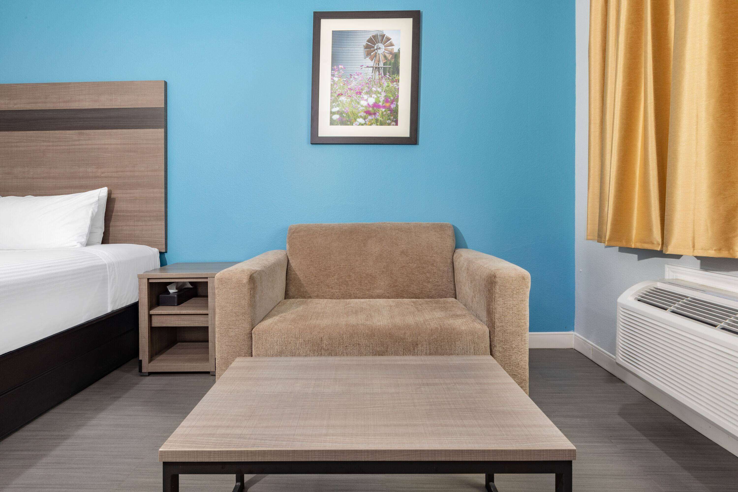 A hotel room with a bed, a small table, a chair, a nightstand, a framed picture on the wall, and a window with curtains.