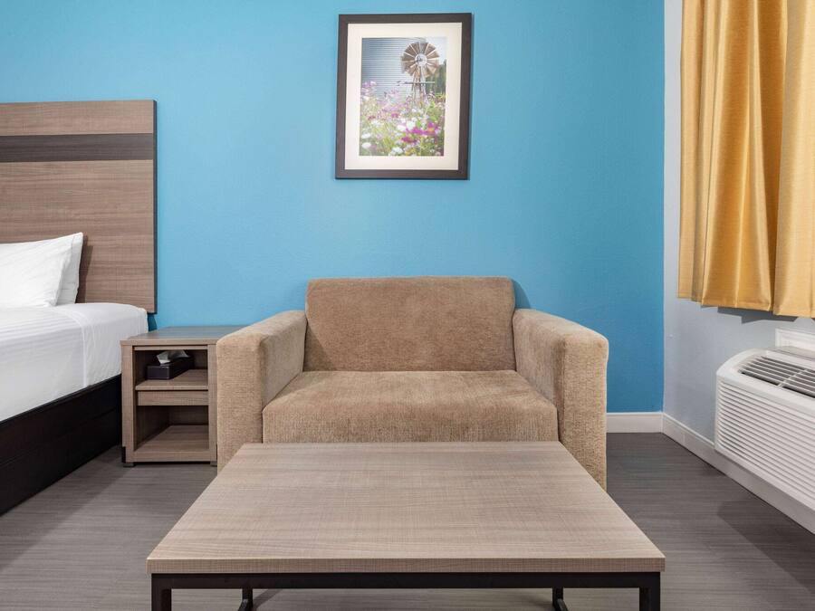 A hotel room with a bed, a small table, a chair, a nightstand, a framed picture on the wall, and a window with curtains.
