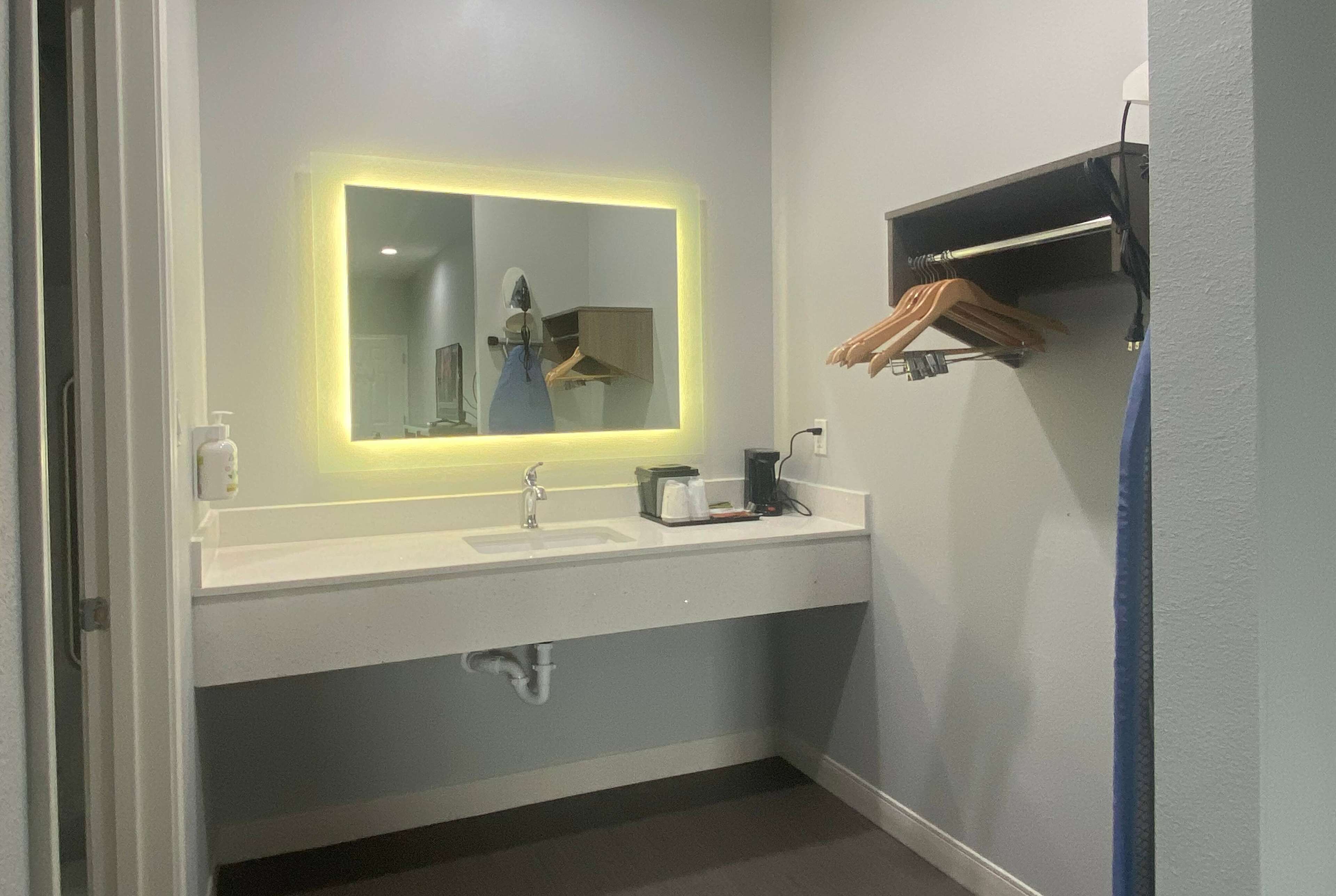 Une salle de bain avec un lavabo blanc, un miroir éclairé d’une lumière jaune, un porte-serviettes et une étagère avec des vêtements suspendus.