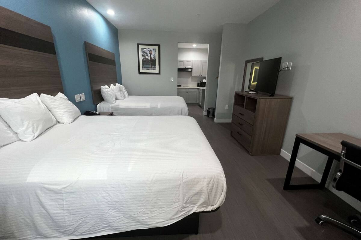 Ein Hotelzimmer mit zwei Betten, einem Fernseher, einem Schreibtisch und einer kleinen Küche im Hintergrund.