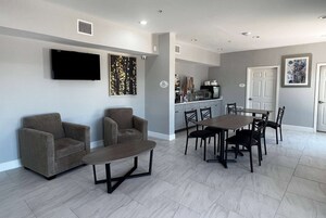 Una sala de estar con dos sofás, una mesa de centro y un televisor montado en la pared. Hay una zona de comedor con una mesa y sillas. Se observa una cocina al fondo, con un refrigerador y un microondas.